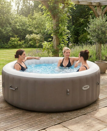Trois femmes profitant d'un spa gonflable rond et gris installé sur une terrasse en bois entourée d'un jardin verdoyant.