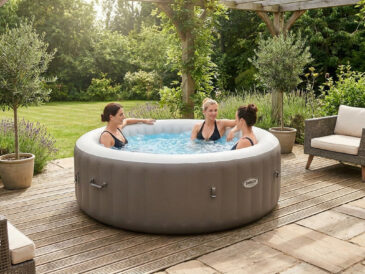 Trois femmes profitant d'un spa gonflable rond et gris installé sur une terrasse en bois entourée d'un jardin verdoyant.