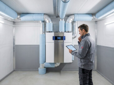 Un technicien vérifie l'installation d'une VMC double flux connectée avec une tablette dans un local technique.